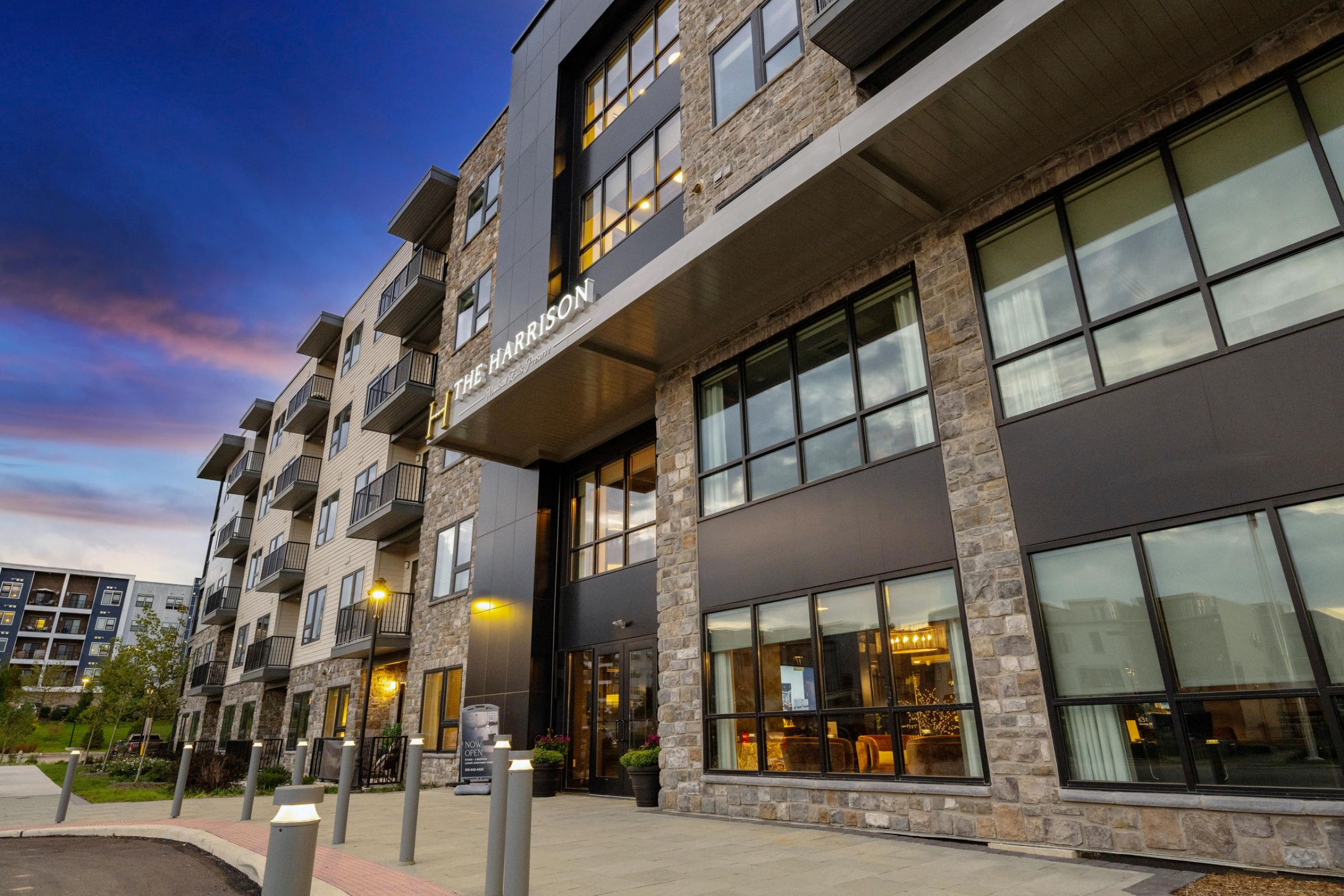 Modern apartment building with large windows and stone facade at sunset, lights glowing inside and outside.