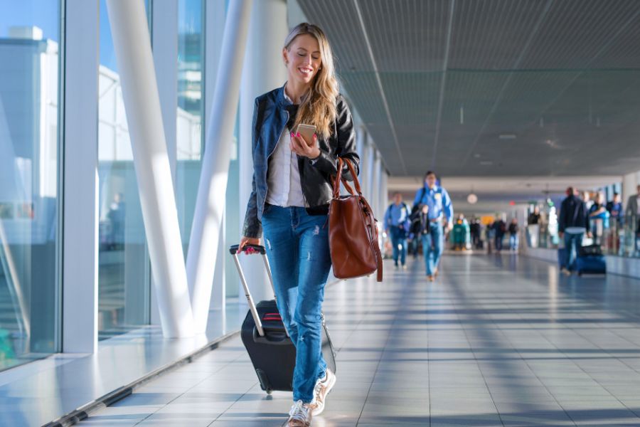Smiling woman walks through an airport with a suitcase, looking at her phone and carrying a brown bag.