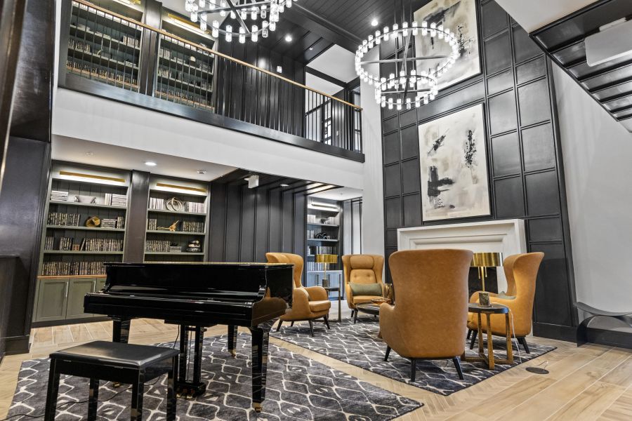Modern library lounge with a grand piano, orange chairs, bookshelves, chandeliers, and abstract art.