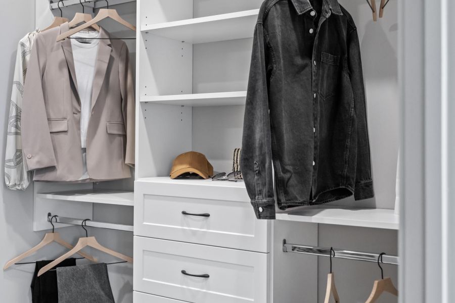 Modern walk-in closet with shelves, drawers, and hanging clothes including jackets, pants, and a brown cap.