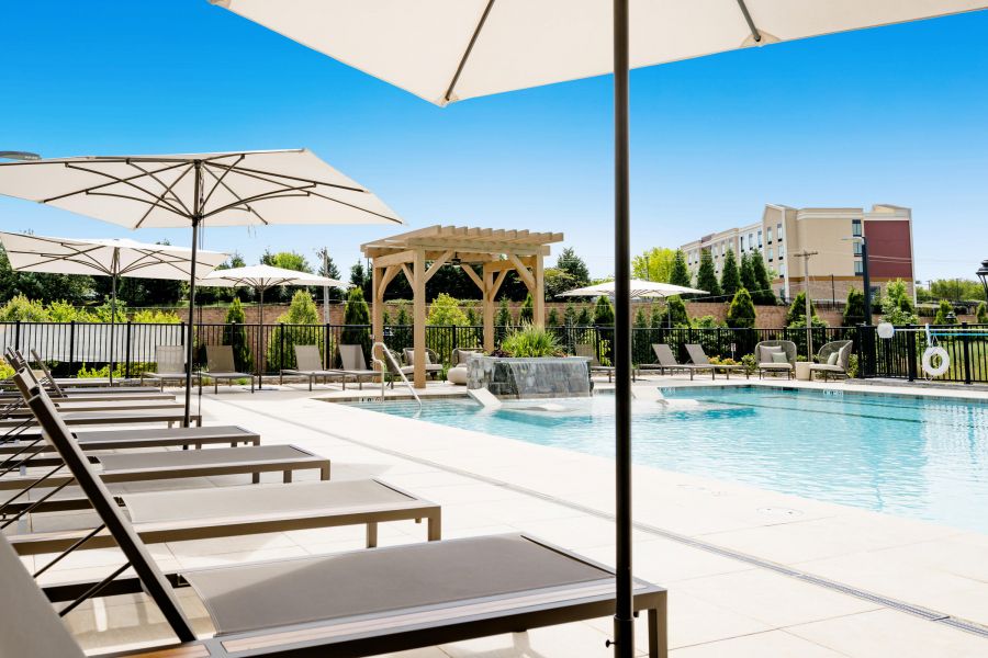 Outdoor pool area with lounge chairs, umbrellas, a pergola, and clear blue sky on a sunny day.