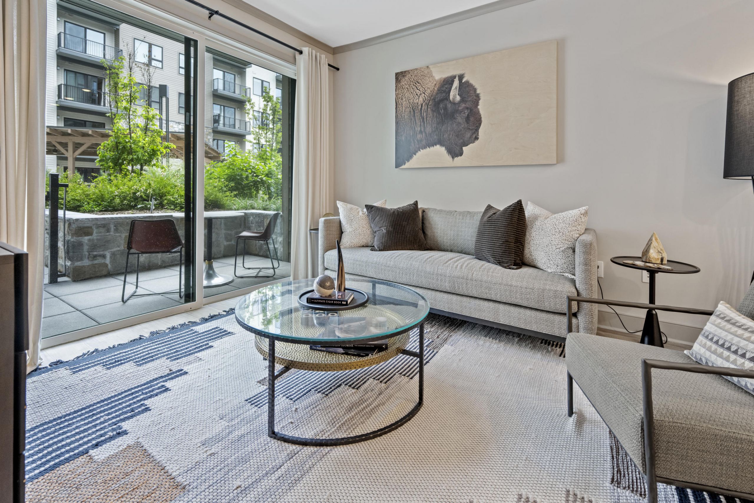 Modern living room with gray sofa, glass coffee table, and large sliding doors leading to a patio with chairs.