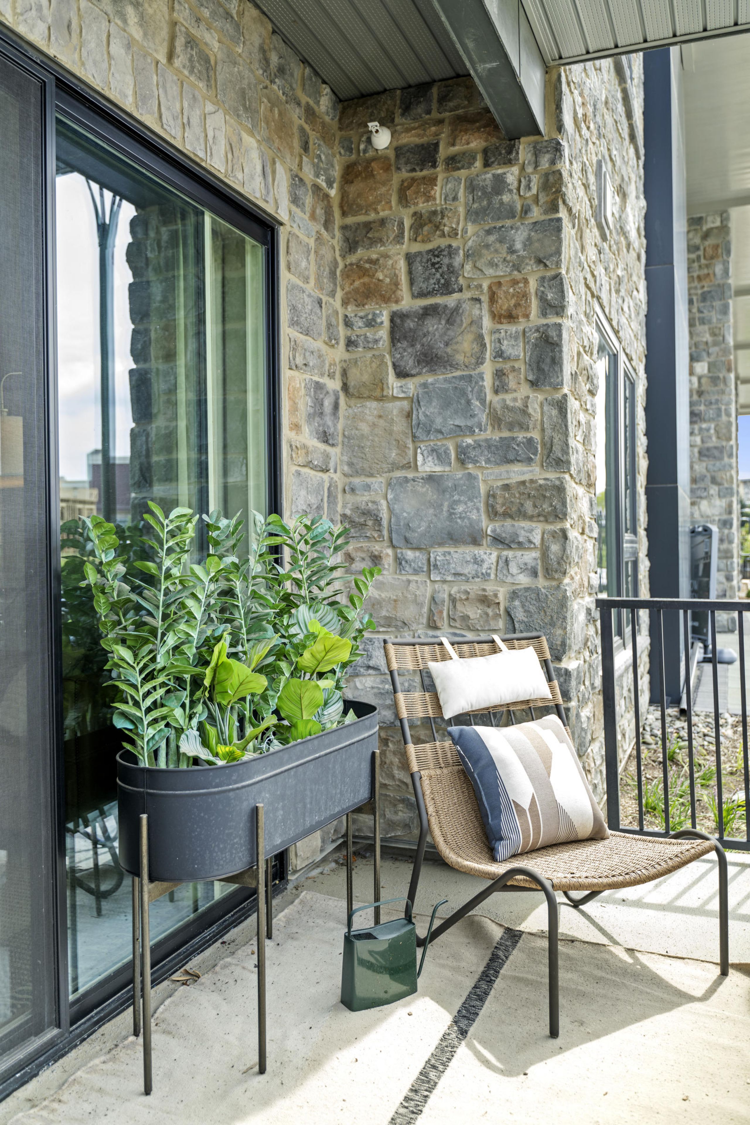Modern patio with a potted green plant, woven chair, and pillows beside a glass door and stone wall.
