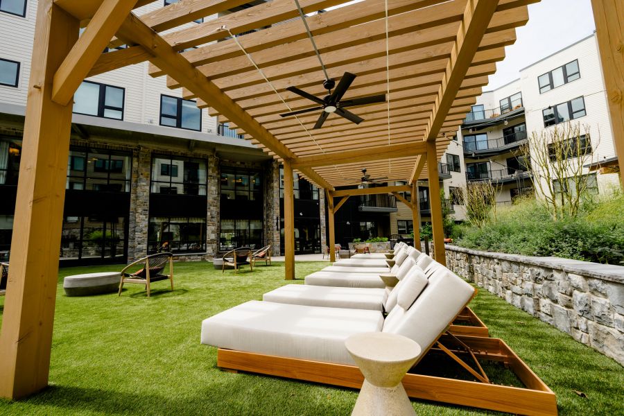 Outdoor lounge chairs under a wooden pergola on a grassy patio, with apartments and greenery in the background.