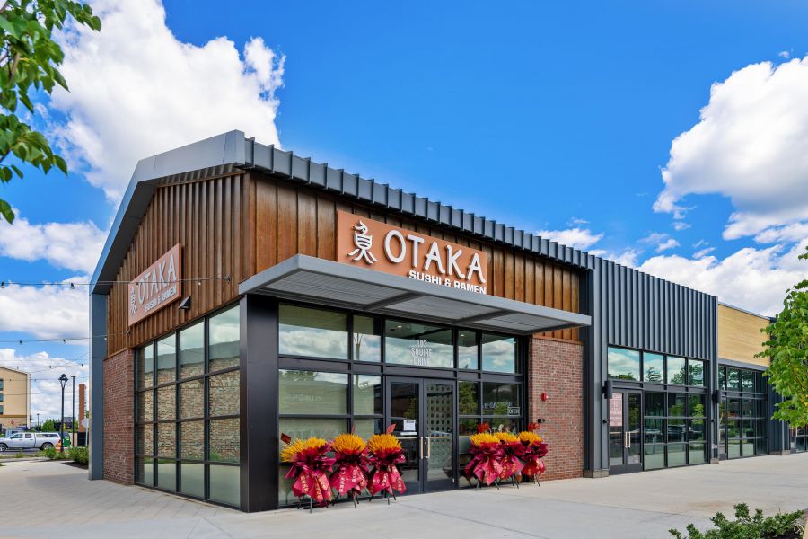 Outdoor shopping plaza with green lawn, colorful chairs, and modern storefronts under a sunny, partly cloudy sky.