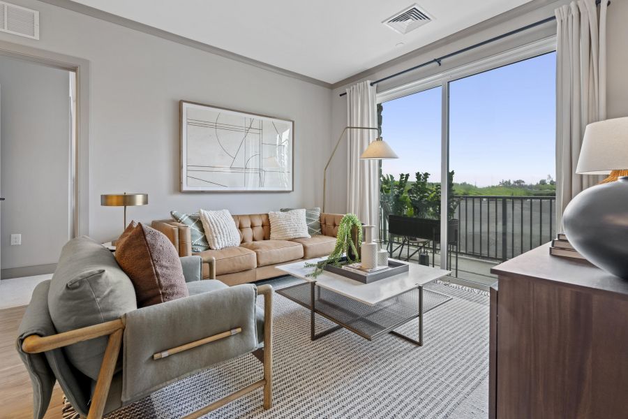 Modern living room with a tan sofa, armchair, coffee table, large art, and sliding glass doors to a balcony.