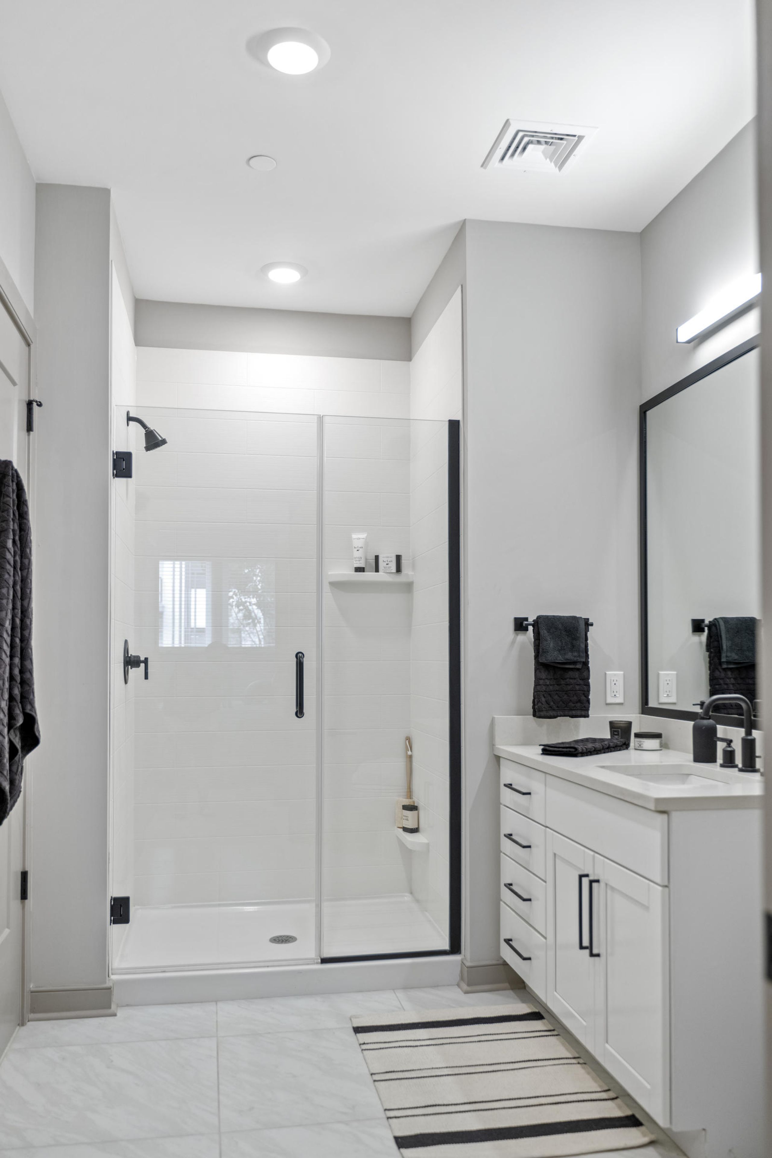 Modern bathroom with a glass shower, white cabinets, a striped rug, and black towel accents.
