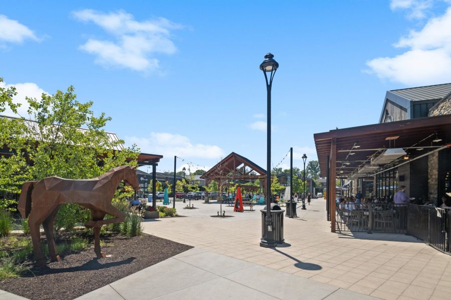 Outdoor shopping and dining area with a metal horse sculpture, trees, and people seated at restaurant tables.