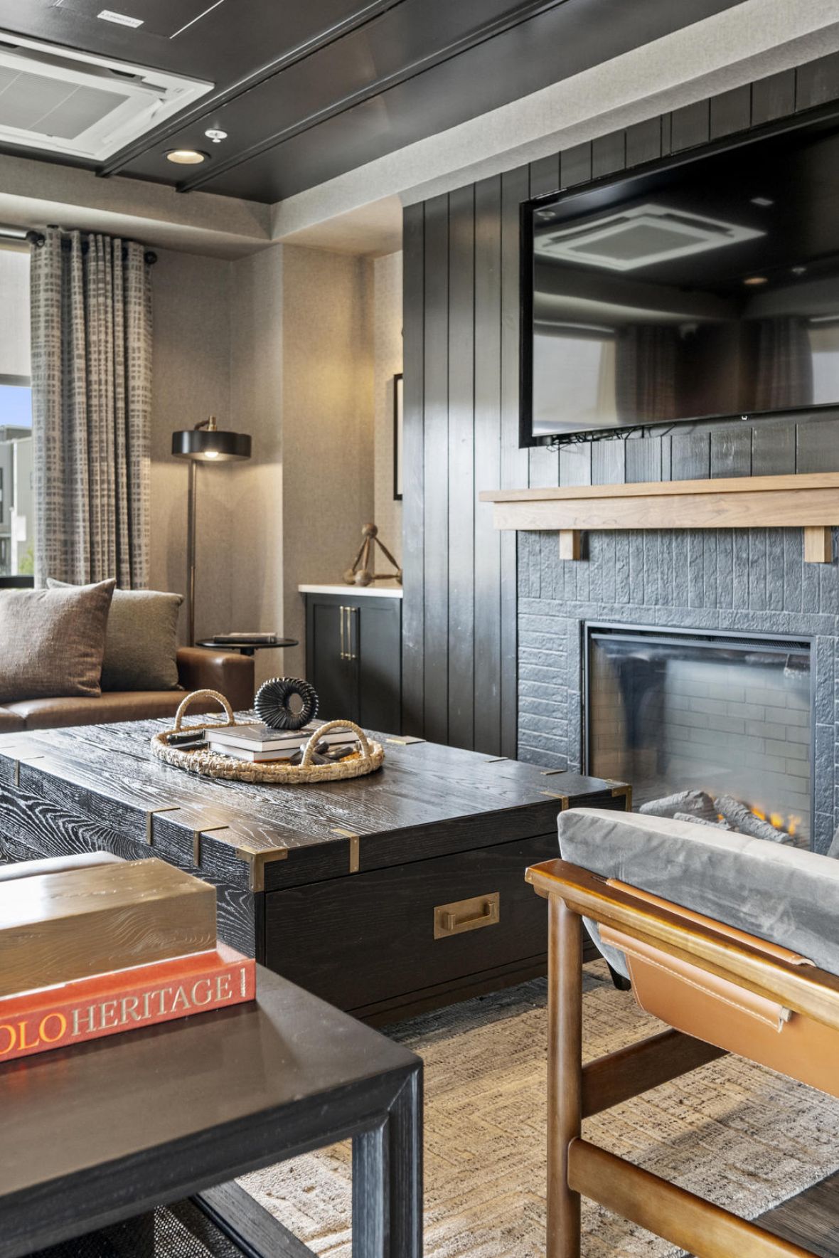 Modern living room with dark walls, fireplace, large TV, sofa, armchair, and window with city view.