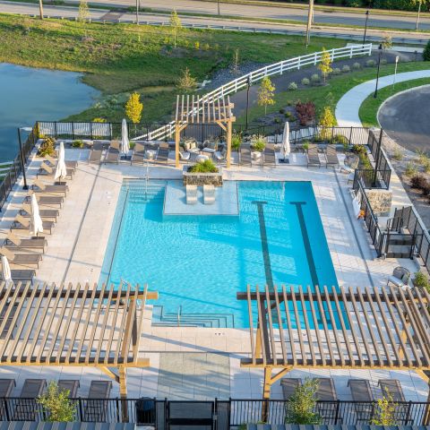 Aerial view of a modern outdoor swimming pool area with lounge chairs, pergolas, and parked cars nearby.
