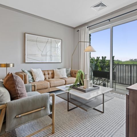 Modern living room with a tan sofa, armchair, coffee table, large art, and sliding glass doors to a balcony.