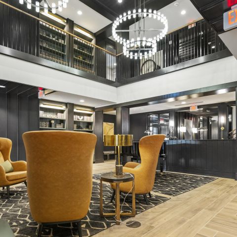Modern lounge with mustard armchairs, black accents, chandeliers, and bookshelves under a mezzanine.