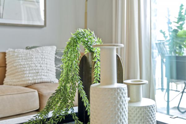 Modern living room with vases, books, and greenery on a tray; sofa, lamp, and window in the background.