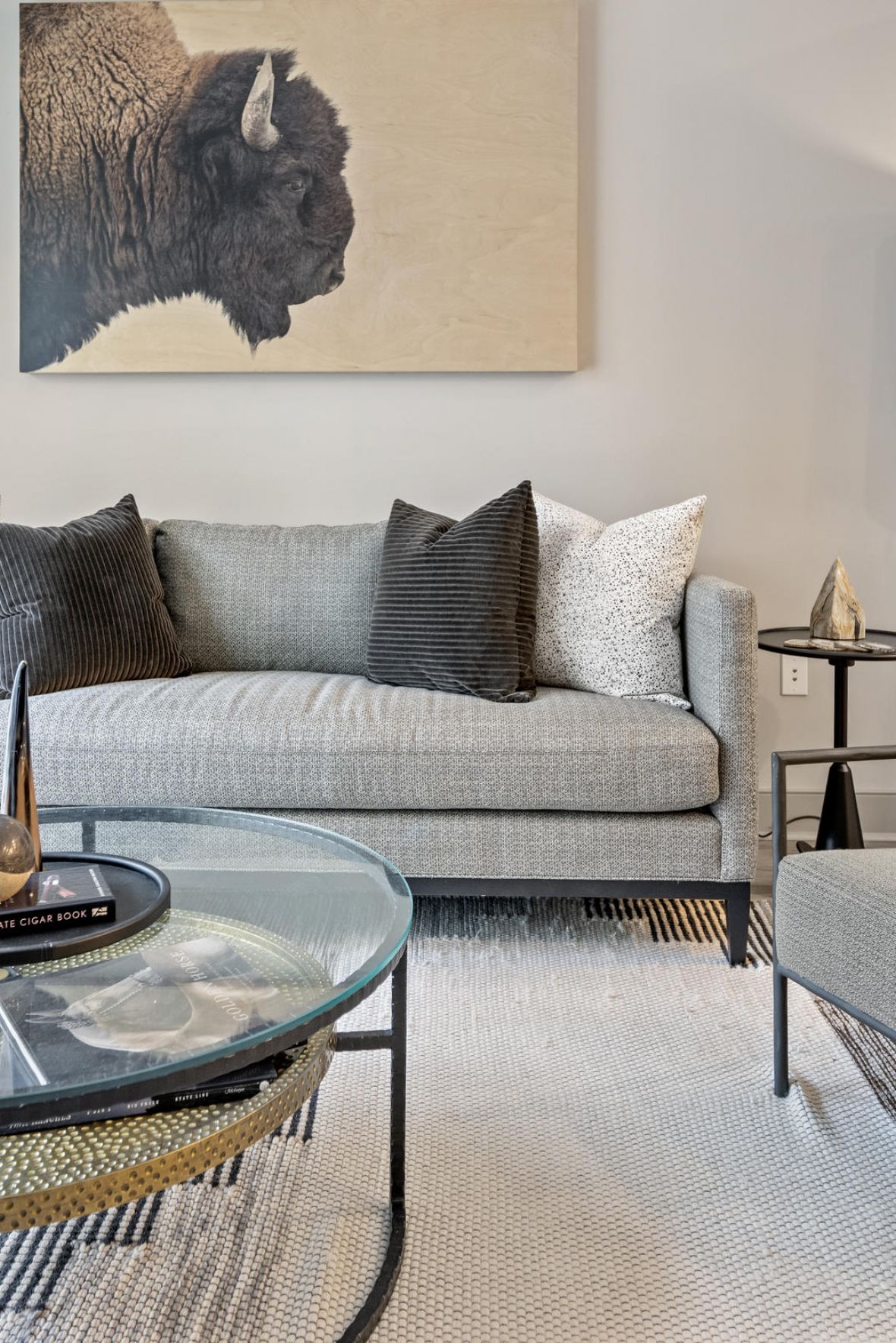 Modern living room with gray sofa, armchair, glass coffee table, large lamp, and bison artwork on the wall.
