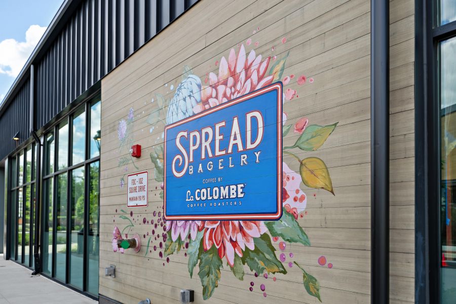 Colorful "Spread Bagelry" mural with flowers on an exterior wall beside large windows at a modern building.