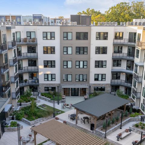 Modern apartment complex courtyard with greenery, seating areas, and pergolas, surrounded by multi-story buildings.