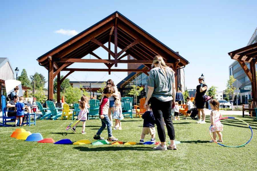 Children and adults play on grass with colorful toys and hoops in front of a wooden pavilion on a sunny day.
