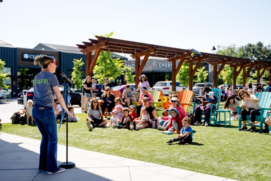 A woman speaks at a microphone to a group of children and adults seated outdoors on a sunny day.