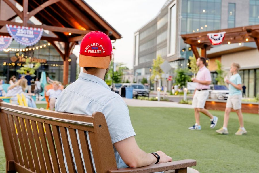 A man in a red hat sits on a bench in a grassy outdoor area with people walking and buildings in the background.