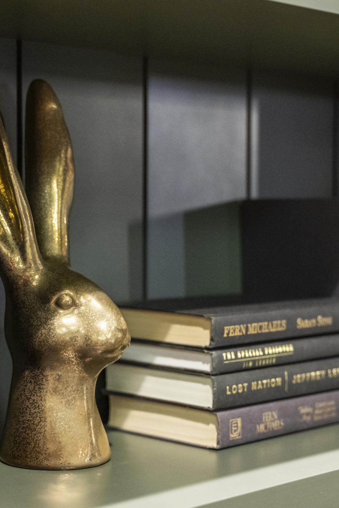 A gold rabbit head sculpture sits on a bookshelf next to neatly arranged dark-colored books.