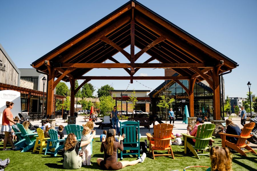 People sit in colorful chairs under a wooden pavilion at an outdoor event on a sunny day.