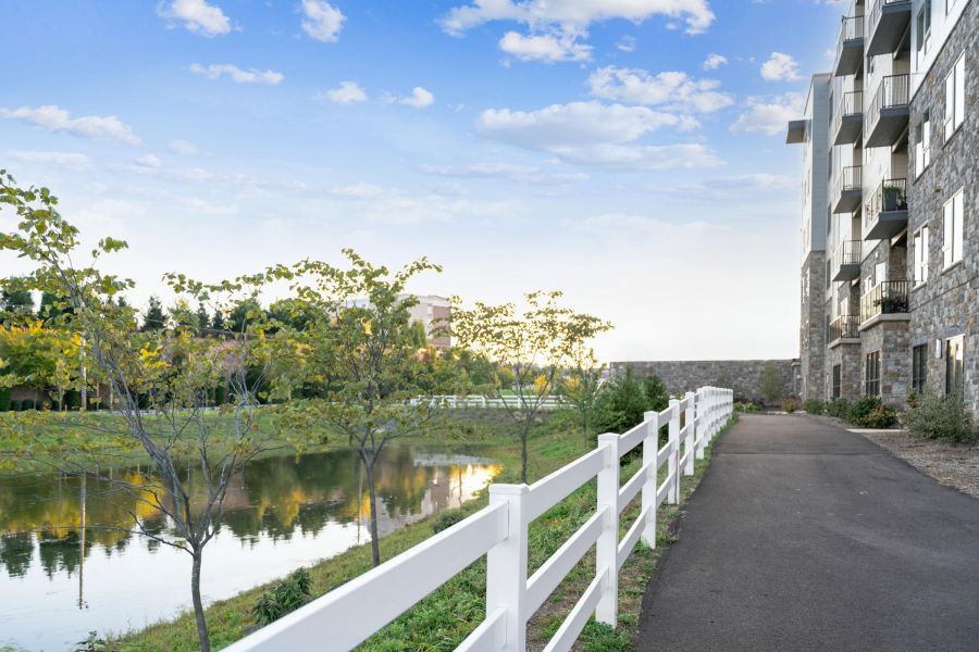Paved path with white fence beside a pond and modern apartment building under a blue sky.
