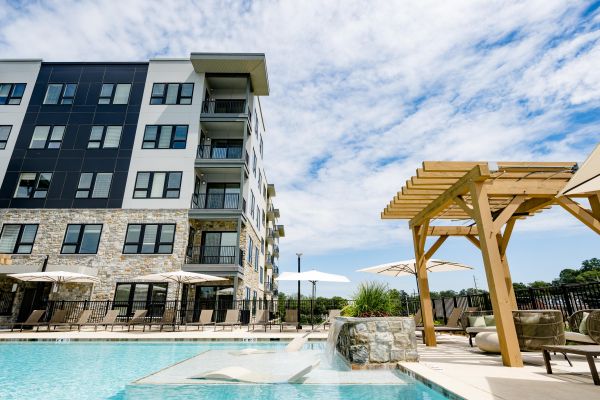 Modern apartment building with a pool, lounge chairs, umbrellas, and a wooden pergola under a partly cloudy sky.