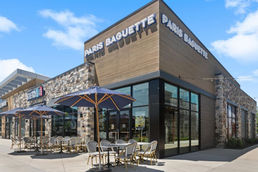 Paris Baguette bakery exterior with outdoor seating, umbrellas, stone walls, and clear blue sky.